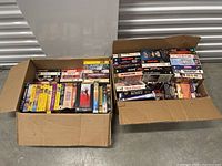 Two open cardboard boxes filled with assorted VHS movie tapes showing a variety of movie titles stacked inside.