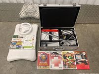 Full view of the gaming bundle showing Wii Fit balance board with steering wheel, four Wii games, and the carrying case open containing the console and cables.