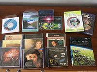 Photo of assorted DVDs and CDs displayed on a wooden surface showing titles including Planet Earth Complete Series DVD, multiple music CDs including Cecilia Bartoli, Andrea Bocelli, Jose Carreras, Andrew Lloyd Webber, Introduction to French CDs, and Bill Cosby spoken word CD.