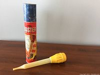 Photo showing the kitchen baster with original cylindrical packaging standing upright on a wooden surface.
