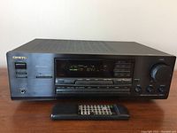 Front view of Onkyo TX-8511 receiver with remote control placed in front, showing display lit up at FM 104.2 MHz and control buttons.