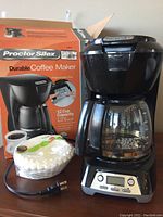 Black Proctor Silex Durable Coffee Maker with glass carafe, digital clock and buttons on the base, original box and coffee filters shown.