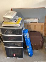 Four drawer plastic storage bin, art supplies, drawing pads, cardboard box, and a rolling brown art bag next to a table easel.