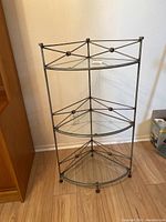 Front view of metal and glass corner shelf with three glass tiers and decorative metal crossbars.