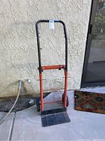 Full view of metal moving dolly standing upright on pavement against a beige stucco wall. Shows handle, frame, and platform base with visible rust and paint wear.