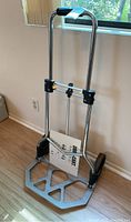Full view of the Duracart dolly hand truck standing upright next to a wall, showing metal frame, black grips, and instruction manual attached.