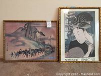 Photo of two framed art pieces side by side on the floor against a wall. The left piece is a watercolor-style painting of travelers and mountains, the right piece is an Asian geisha styled poster print with text.