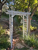 Full view of white wooden garden arbor installed outside in a garden setting with plants, stone steps, and a bird feeder hanging inside the arbor.