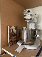 White KitchenAid stand mixer with bowl, attachments, and manual on shelf.