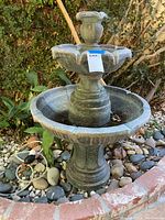 Front and angled view of two-tier stone outdoor fountain with visible scalloped edges and textured green-gray finish placed in garden setting.