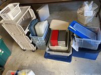 Wide view showing plastic bins with lids, buckets, rolling storage cart, and reusable bags stacked on floor.
