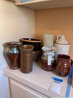 Seven pottery items arranged on a white shelf including vases, pots, bowls, and jugs with varied earthy colors and finishes.