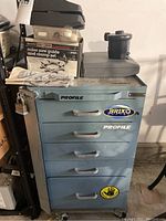Blue Profile metal tool box on wheels with five drawers, stickers on front, with small black/gray device and Sears Craftsman miter saw guide and clamp set box on top plus gloves.