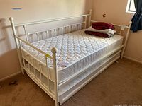 White metal trundle bed frame with two mattresses, one on the pull-out trundle below. Linens and bedding folded and stacked on top mattress.