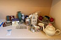 Wide view showing the assortment of Wedgwood items, ceramic vases, faux fruit on tray, teapot set, and cutlery rests on a white shelf.