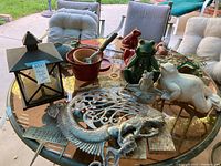 Wide view of all yard art items arranged on glass outdoor table including frogs, fish, mermaid metal ornament, candle house, garden pot, and hand rake.