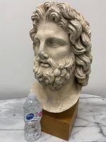 Front view of large ancient Greek male head bust on teak base next to a water bottle for scale.