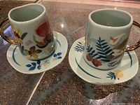 Pair of ceramic Disney Mary Blair floral tea cups and saucers with gold handles and colorful flower designs, shown on clear surface.