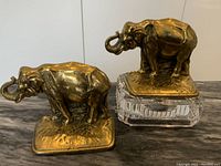 Pair of heavy vintage brass elephant book ends with one elephant on a textured brass base and the other on a glass block stand (stand not included).
