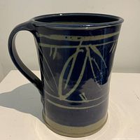 Dark blue pottery mug with light blue leaf motif and a large handle, shown from an angle to highlight the hand-painted design and ceramic finish.