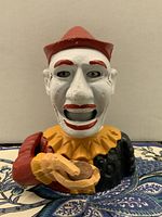 Front view of vintage painted iron clown penny bank holding a coin in its hand, showing face, hat, collar, and outfit colors.