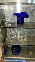 Full shelf view showing all items including origami bird, blue art glass vase, clear crystal wine decanter, yellow dish, and another blue vase.
