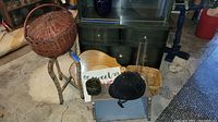View of two round woven baskets, wooden cutting boards, black equestrian helmet, small glass vase, small woven basket, and folding metal stand on concrete floor.