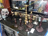Photo shows the grouping of six wooden candlesticks, three brass candlesticks, one pewter candlestick with patina, and three clear acrylic bobeches with hanging acrylic prisms arranged on a dark surface in front of a mirror.