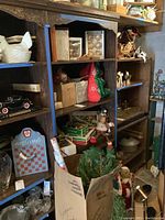 Shelf with various vintage Christmas decorations, boxes, a white ceramic hen, and artificial greenery in a box below.