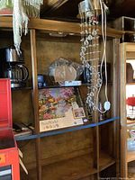 Wooden shelving unit holding various items including a PaintWorks paint by number kit, a round mirror, vintage leather carrying case, and a purse lamp hanging from the top shelf with beaded decoration.