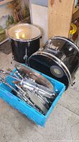 Blue crate with disassembled drum hardware and stands next to two black drums on the floor