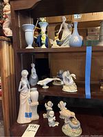 Shelf display of various figurines and vases including goose marked Lladro, figurines marked Hummelworks, and several blue vases.