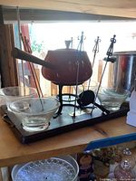 Full view of the fondue set with pot, forks, and glass bowls on wooden shelf near window, showing red fondue pot with wooden handle, black iron stand, clear glass bowls, and fondue forks with colorful handles