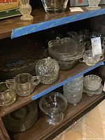 Shelf with glass creamers, sugar dishes, lidded candy dishes, lunch trays, plates, platters, trays in clear vintage glass with textured and patterned designs.