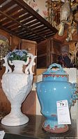 Both the white porcelain vase and the blue glazed pottery urn shown side by side on a shelf with background wooden cabinets and vintage decor.