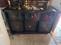Front view of black wooden Mid Century Modern style buffet cabinet with a drawer and three sliding doors, placed indoors on concrete floor and surrounded by brass and glass candlesticks on top.
