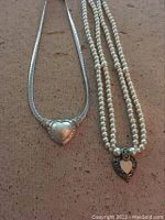 Two necklaces laid out on a neutral fabric background. One necklace has a silver smooth heart pendant with a flat silver chain, the other a double strand of faux pearls with a decorative heart pendant.
