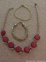 All three jewelry pieces displayed on fabric background: one necklace with pink round stones and two gold-tone chain bracelets.