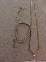 Photo showing the pair of gold-tone earrings, gold-tone necklace with v-shaped clasp, and matching gold-tone bracelet with bead accents placed on a beige textured fabric.