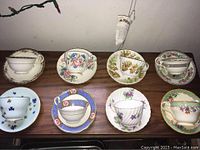 Eight Aynsley teacups and saucers laid out on a wooden surface, showing the full set and variety of designs including floral patterns and gold detailing.