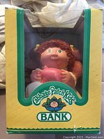 Front-facing view of the Cabbage Patch Kids bank figure inside a yellow and green box with clear window showing the doll holding a pink piggy bank.