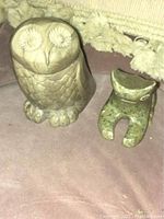 Photo showing both soapstone animal carvings: owl with detailed feather etching and large eyes, frog with a smooth abstract shape, both resting on a suede surface.