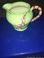 Front view of the green ceramic mini pitcher with pink foxglove flower and bamboo-style handle on blue surface.