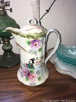 Front and side views of the hand painted antique Japanese cocoa pot on wooden surface with adjacent lamp and dishware.