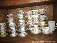 Multiple stacks of vintage tea cups and demitasse cups with matching saucers, floral and decorative patterns, arranged on a wooden shelf.