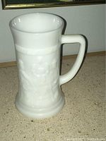 Full view of the milk glass stein mug showing the handle, flared lip, and embossed relief scene on the body.