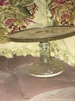 Side view of vintage glass cake stand showing embossed floral pattern on pedestal base and textured glass plate, placed on pink tufted fabric with floral background.