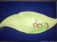 Top view of light green leaf-shaped ceramic celery dish with embossed veins and sculpted pink flowers near the wider end, on blue surface.