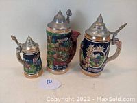 Photo showing the set of three German beer steins with pewter lids and raised detailed designs placed on a white surface