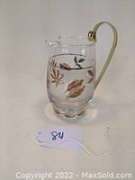 Small clear glass pitcher with gold leaf and foliage designs, gold-tone curved handle visible, placed on white surface with ID tag '84'.